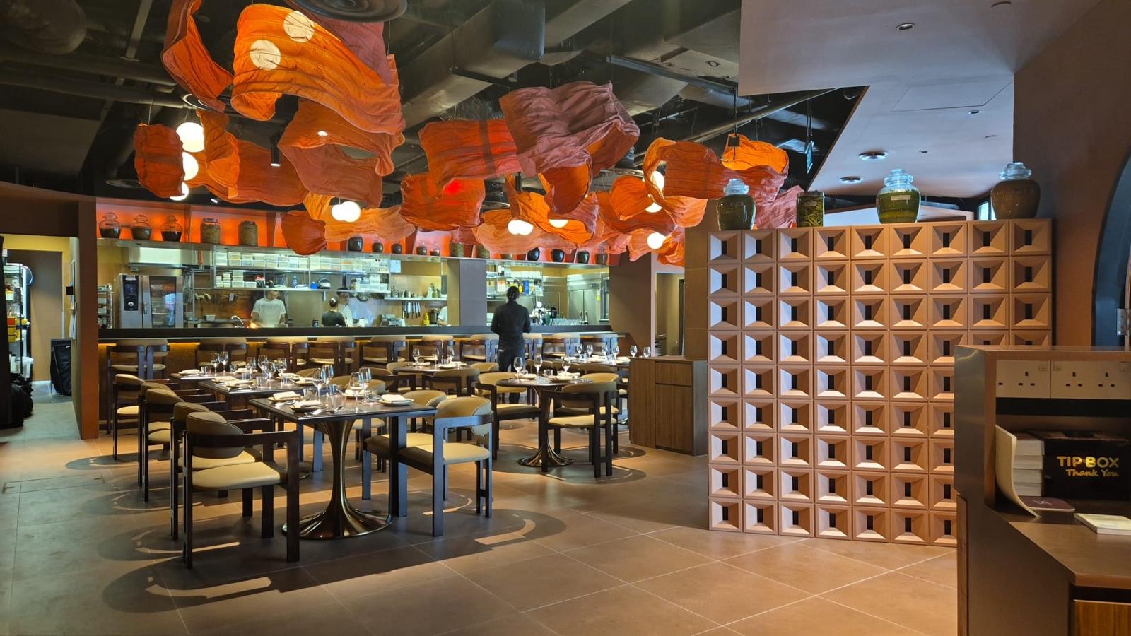 LIORA restaurant interior with orange pendant lighting, dining tables, an open kitchen and a brick screen